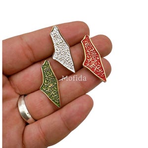 May include: Three enamel pins in the shape of a map, each with a gold border. One is white with black script, one is red with white script, and one is green with gold script. The pins are displayed on a white background.