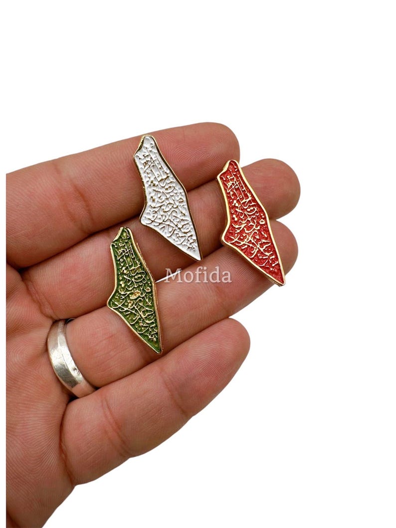 May include: Three enamel pins in the shape of a map, each with a gold border. One is white with black script, one is red with white script, and one is green with gold script. The pins are displayed on a white background.