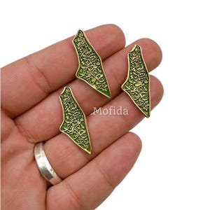 May include: Three gold-colored, leaf-shaped pendants with intricate green detailing and Arabic script. The pendants are displayed on a hand, with a silver ring visible. The word "Mofida" is printed on the image.