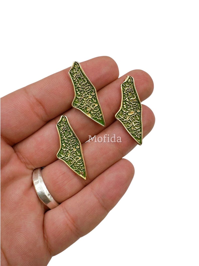May include: Three gold-colored, leaf-shaped pendants with intricate green detailing and Arabic script. The pendants are displayed on a hand, with a silver ring visible. The word "Mofida" is printed on the image.