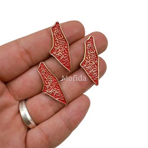 May include: Three red and gold-colored pendants in the shape of a map. The pendants have intricate gold detailing and are held in a hand. The word "Mofida" is visible on the image.