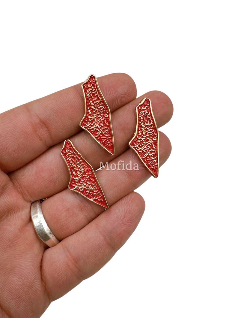 May include: Three red and gold-colored pendants in the shape of a map. The pendants have intricate gold detailing and are held in a hand. The word "Mofida" is visible on the image.
