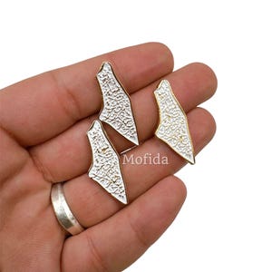 May include: Three white and gold-colored metal charms in a unique shape, held in a hand. The charms have a textured white surface and a gold-colored border. The word "Mofida" is visible on the image.