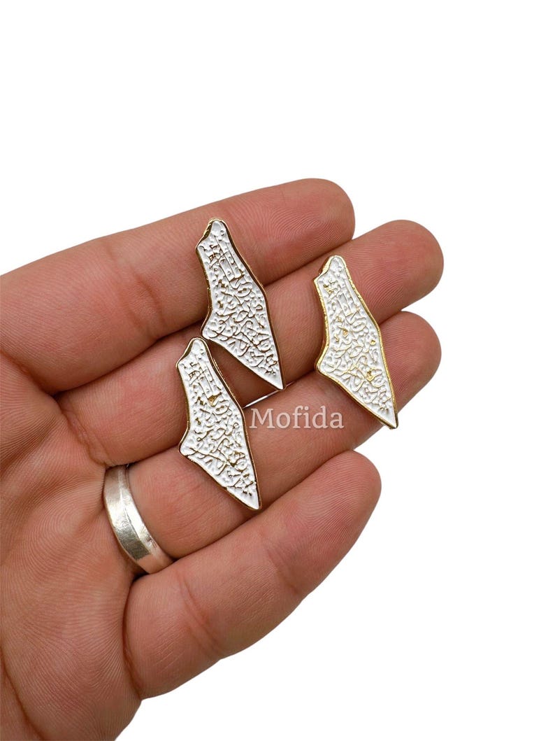 May include: Three white and gold-colored metal charms in a unique shape, held in a hand. The charms have a textured white surface and a gold-colored border. The word "Mofida" is visible on the image.