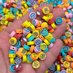 May include: A collection of colorful, round beads with various cartoon character designs. The beads are primarily yellow, pink, blue, and orange, with detailed illustrations. These beads are ideal for crafting and jewelry making.