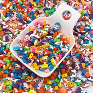 May include: A collection of colorful rainbow-shaped beads in various colors, including orange, yellow, pink, blue, and green. The beads are scattered around a white scoop, creating a vibrant and playful display. Ideal for jewelry making or craft projects.