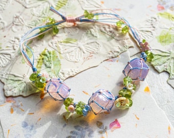 May include: A delicate necklace with pink and purple glass beads wrapped in blue and pink string. The necklace is accented with green and white flower-shaped beads.