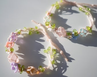 May include: Two delicate bracelets with pink, green, and blue crystals and crocheted flowers. The bracelets are made with white string and have a bohemian style.