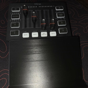 May include: A black audio mixer with the brand name "fifine" printed on the top. It features multiple sliders, buttons, and input/output options. A black stand is in the foreground. The mixer is designed for audio recording and mixing.