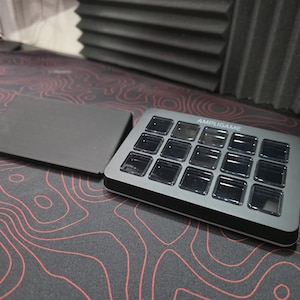 May include: A gray Ampligame controller with 15 square buttons and a separate gray stand. The controller is on a black and red topographic map-patterned surface. The word AMPLIGAME is printed on the controller.