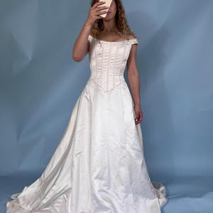 May include: A white satin wedding dress with a fitted bodice and a flowing skirt. The dress features a sweetheart neckline and off-the-shoulder sleeves. The skirt has a long train that pools on the floor. The dress is a classic bridal style.