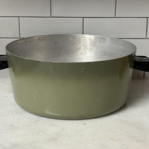 May include: A round cooking pot with a matte olive green exterior and a metallic silver interior. The pot features two black rectangular handles on opposite sides. The exterior surface shows minor surface scratches and signs of previous use.