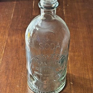 Pode incluir: Garrafa de refrigerante de vidro transparente com texto em relevo. A garrafa tem um corpo arredondado e um gargalo estreito. O texto inclui a palavra "SODA" e o nome "E. GAZET MARTIN FRERES & Cie".