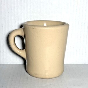 May include: A small, beige ceramic mug with a rounded base and a curved handle. The mug has a simple, classic design, suitable for coffee or tea. The mug is approximately 3 inches tall.
