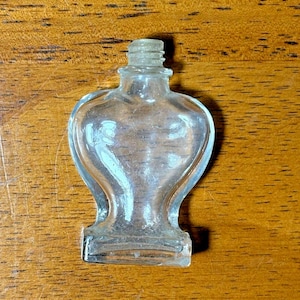 Pode incluir: Um frasco de perfume de vidro transparente e vintage com um corpo em forma de coração e um gargalo roscado. O frasco tem uma base retangular e é exibido em uma superfície de madeira. O frasco está vazio e parece antigo.