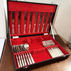 Könnte beinhalten: Ein schwarzer Aufbewahrungskoffer mit leuchtend rotem Samtinnenfutter, der ein Set aus silberfarbenem Besteck enthält. Das Set umfasst acht Messer im Deckel sowie acht Löffel und acht Gabeln mit verzierten Griffen, die im Boden angeordnet sind.