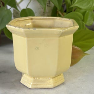 Puede incluir: Una maceta de cerámica de color amarillo pálido con forma octogonal y base de pedestal. La superficie esmaltada presenta sutiles signos de desgaste a lo largo del borde superior, incluyendo pequeñas muescas y decoloración.