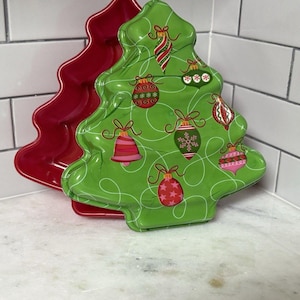 Puede incluir: Dos fuentes con forma de árbol de Navidad. Una es roja, la otra verde y decorada con adornos y espirales blancas. Las fuentes son de un material brillante y están sobre una superficie blanca.