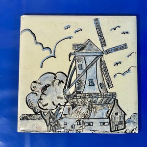 May include: A hand-painted ceramic tile depicting a blue and white windmill with a house in front of it. The tile has a cream background with blue clouds and birds.