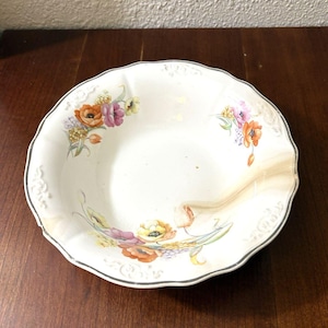 May include: A white, scalloped-edge ceramic bowl with floral designs in orange, pink, yellow, and purple. The bowl has a dark gray trim and a decorative border. The bowl is likely vintage or antique.
