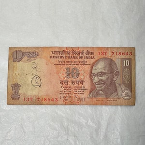 Puede incluir: Un billete indio de diez rupias desgastado. El billete presenta texto en varios idiomas, incluyendo "RESERVE BANK OF INDIA" y "I PROMISE TO PAY THE BEARER THE SUM OF TEN RUPEES." Se ve un retrato de Mahatma Gandhi.