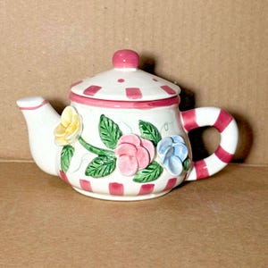 May include: A small white teapot with a pink lid and handle. The teapot is decorated with pink, blue, and yellow flowers and green leaves. The handle is striped pink and white.