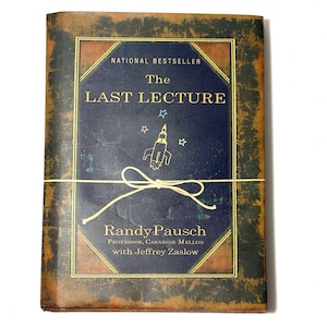 Könnte beinhalten: Ein gebundenes Buch mit dem Titel "The Last Lecture" von Randy Pausch. Das Buch hat einen marineblauen Einband mit goldener Schrift und einer Raketenillustration. Eine Schnur ist um das Buch gebunden.