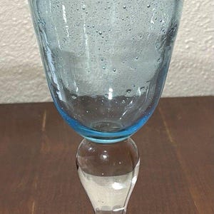 May include: A clear glass goblet with a light blue bowl and a clear stem and base. The bowl has a flared shape and is filled with tiny bubbles. The goblet sits on a dark brown wooden surface.