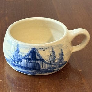 May include: A small, off-white ceramic mug with a rounded body and a handle. The mug features a hand-painted blue landscape scene of a barn and trees. The interior of the mug is a creamy color.