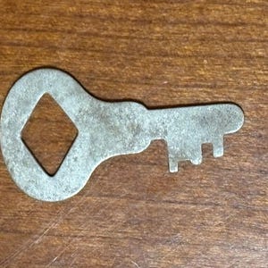 Könnte beinhalten: Ein Schlüssel im Vintage-Stil aus mattem, silberfarbenem Metall mit einer rautenförmigen Öffnung im Kopf. Der Schlüssel hat ein abgenutztes, gealtertes Aussehen mit sichtbaren Zähnen. Das Design deutet auf ein antikes oder dekoratives Objekt hin.