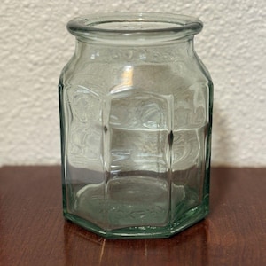 May include: A clear, hexagonal glass jar with a wide mouth and a thick, rounded rim. The jar features six vertical panels and a flat base, constructed from transparent glass with a slight green tint, designed for storage or decorative purposes.