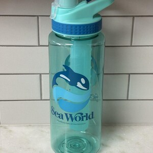 Pode incluir: Uma garrafa de água de plástico translúcido em tom verde-azulado com um gráfico de uma orca em azul escuro e azul claro acima do texto Sea World e SeaWorld Parks & Entertainment. A garrafa tem um colar texturizado azul escuro e tampa.