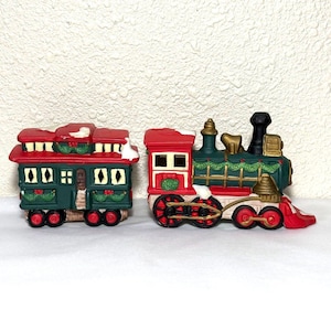May include: A ceramic Christmas train set with a green and red engine and a green and red passenger car. The train has a white snow-covered roof and green garland decorations.