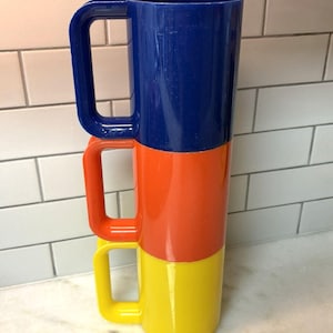 May include: A stack of three cylindrical mugs with square-shaped handles. The top mug is dark blue, the middle mug is bright orange, and the bottom mug is yellow. Each mug features a smooth, glossy finish and a uniform, straight-sided design.