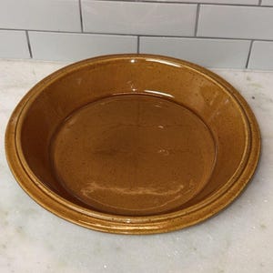 Può includere: Una tortiera rotonda in ceramica marrone con finitura maculata. Il piatto ha un bordo leggermente rialzato e un interno liscio e piatto. La tortiera è progettata per cuocere e servire torte.