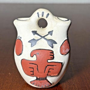 May include: A small, off-white ceramic vase with a handle and a hole. The vase is decorated with a red bird design, black crossed arrows, and red circular accents. The vase is approximately 4 inches tall.