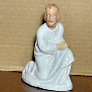 Puede incluir: Una figura de cerámica de una figura arrodillada con una túnica blanca. La figura tiene piel clara, cabello castaño rojizo y barba. La figura está sobre una base blanca.