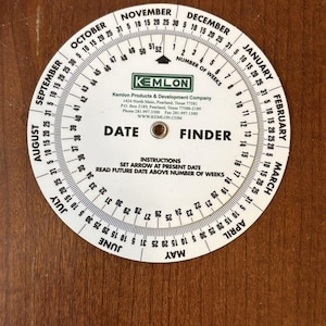 May include: A white Kemlon Date Finder with rotating discs, featuring months, dates, and week numbers. The device includes the Kemlon Products & Development Company logo and instructions. The background is a wooden surface.