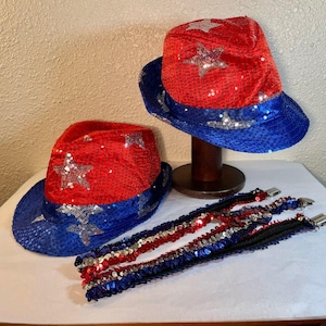 May include: Two fedora hats covered in red, white, and blue sequins featuring silver star patterns. One hat rests on a wooden stand, while the other sits beside it. Three matching sequined suspenders with metal clips are arranged in the foreground.