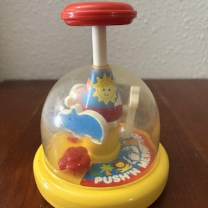 May include: A vintage yellow and clear plastic toy with a red top and a white stem. Inside the clear dome are colorful shapes and the text "PUSH'N MER". The toy is designed for children.