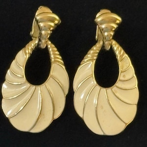 May include: A pair of vintage clip-on earrings. The earrings have a gold-tone metal finish with a cream-coloured enamel inlay. The design features a teardrop shape with a spiral fan design.