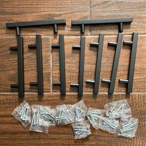 May include: A set of eight matte black cabinet pulls with a T-bar design, each featuring two cylindrical mounting posts. The hardware set includes eight small clear plastic bags, each containing two silver metal mounting screws for installation.