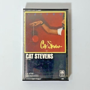 Könnte beinhalten: Eine Vintage-Kassette des Albums "Izitso" von Cat Stevens. Das Cover zeigt ein rotes und gelbes Design mit dem Namen des Künstlers und dem Albumtitel in weißen Buchstaben. Die Kassette hat ein transparentes Gehäuse mit schwarzem Rahmen.