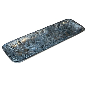 Puede incluir: Una bandeja de servir plateada con un diseño en relieve de una escena tropical. La bandeja es rectangular y tiene un borde ligeramente curvado.