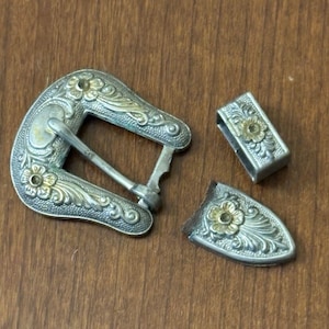 May include: A three-piece silver-tone belt buckle set featuring an engraved floral pattern with gold-tone accents. The set includes a large buckle frame with a prong, a rectangular belt loop, and a pointed belt tip, all decorated with matching floral motifs.