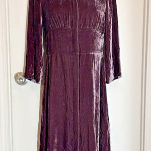 May include: A long-sleeved midi dress in a deep plum purple crushed velvet fabric. The garment features a V-neckline, a fitted bodice with vertical seaming, and a button-down front closure that extends from the neckline to the hemline.