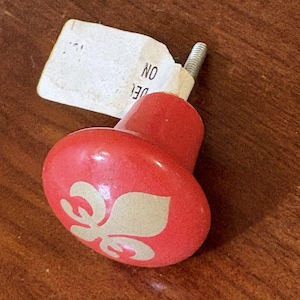 May include: A round, red cabinet knob featuring a white fleur-de-lis design on its face. The knob has a metal screw extending from the back and includes a small, white paper tag with the partially visible text "DEL ON" attached to the screw.