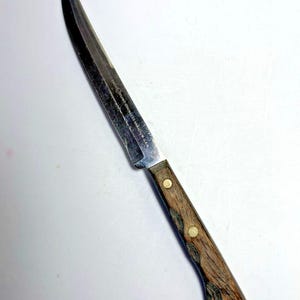 May include: A stainless steel knife with a wooden handle. The blade is long and slender, with a pointed tip. The handle is brown with two brass rivets. The knife has text on the blade.