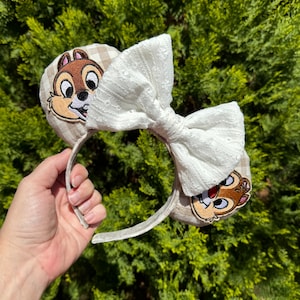 May include: A pair of mouse ears featuring Chip and Dale character patches. The ears have a tan and white checkered pattern and a large white lace bow. The headband is held by a hand against a backdrop of green foliage.