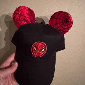 May include: Black baseball cap with red Spider-Man logo and red velvet ears with black spiderweb design. The cap features a curved brim and a circular patch with the Spider-Man mask. A fun accessory for fans.